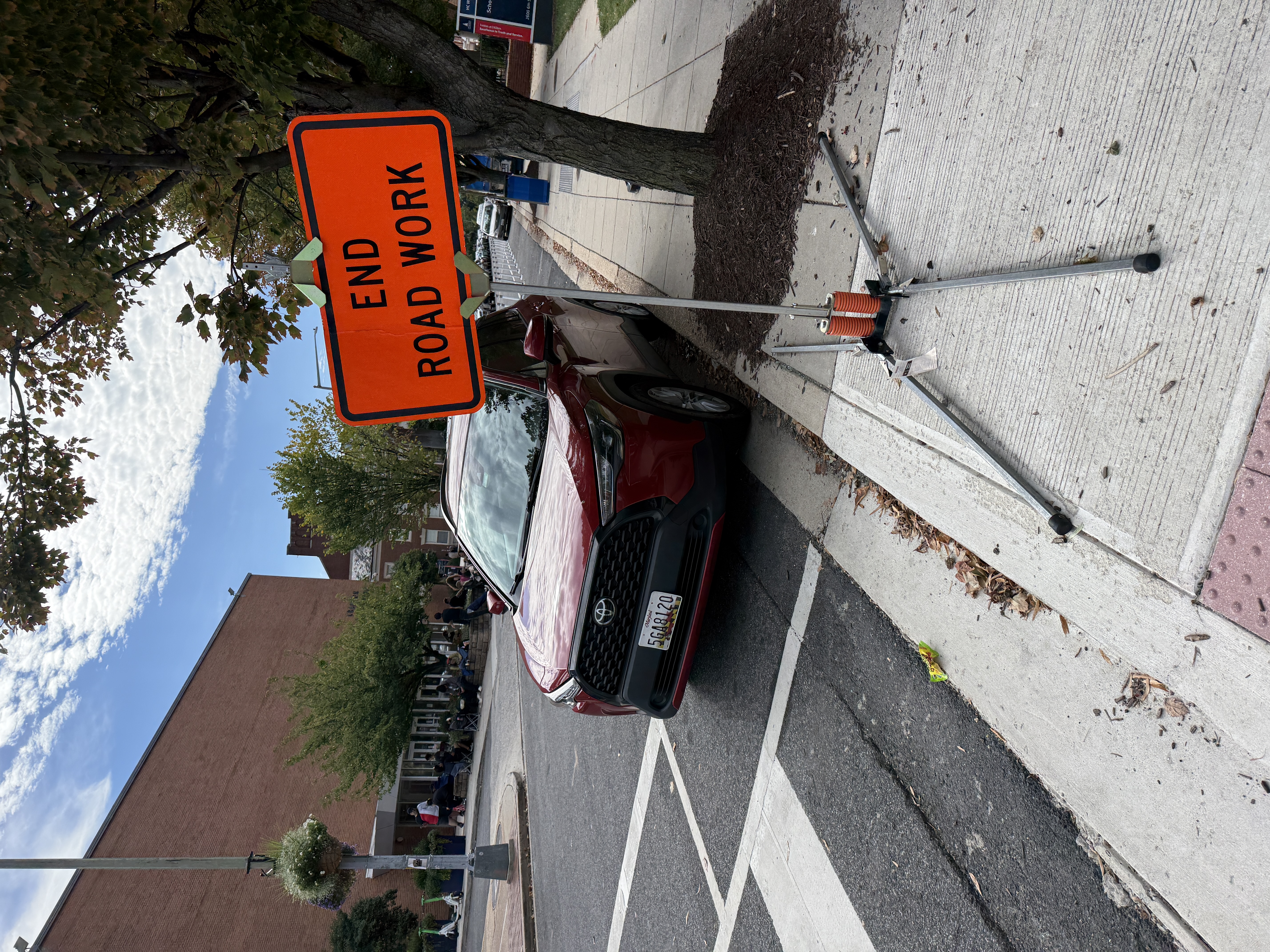 Red sedan parked right up against the crosswalk on the Howard University box office block of 6th St NW at Fairmont. Nothing physically protects the daylighting zone at this corner -- no bollards, no curb extension, no setback marking -- so vehicles park flush to the crosswalk on the same corridor where a Howard student was killed by a speeding driver. The only sign in view is a temporary END ROAD WORK sign.