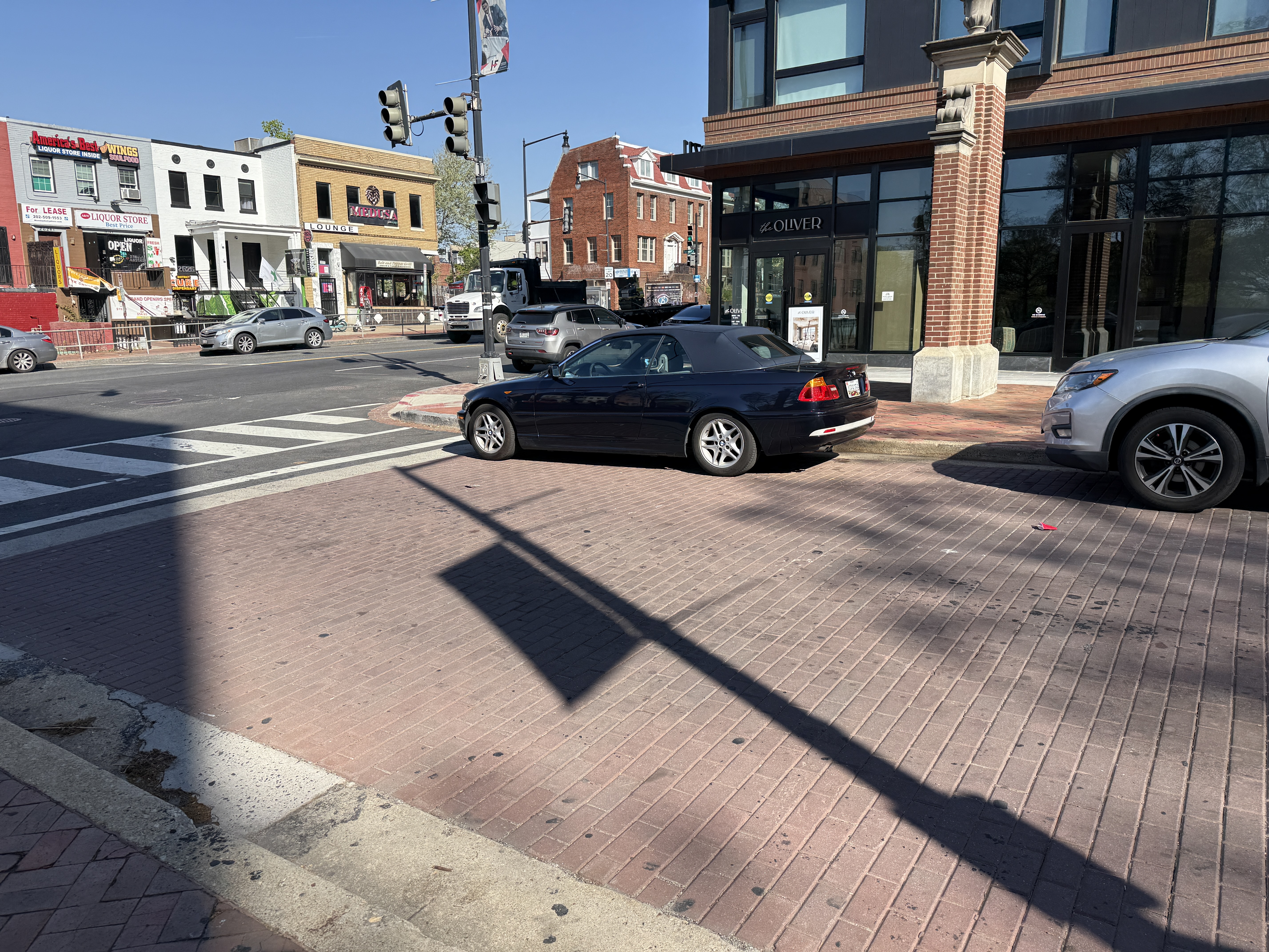 Dark BMW with Maryland plates parked right up against the crosswalk on Georgia Ave NW at Fairmont, near Howard University. No bollard, curb extension, or daylighting marking protects this corner, so vehicles park inside the crosswalk setback on a corridor where a Howard student was killed.