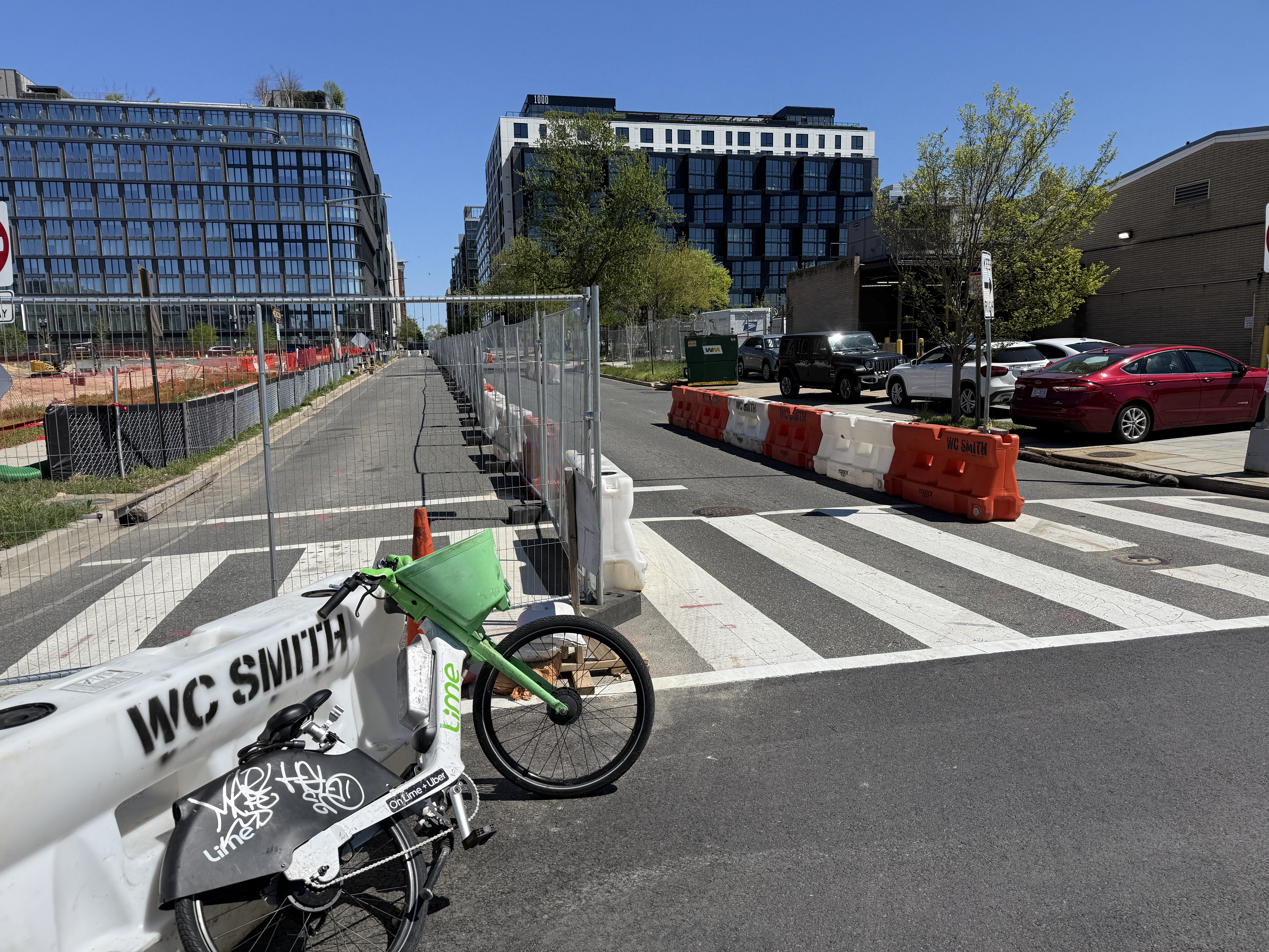 WC Smith construction closed sidewalk with no safe pedestrian accommodation. Cars parked on remaining sidewalk. Abandoned Lime e-bike blocks crosswalk. No wheelchair-accessible path exists on this block -- complete ADA failure and construction permitting violation.