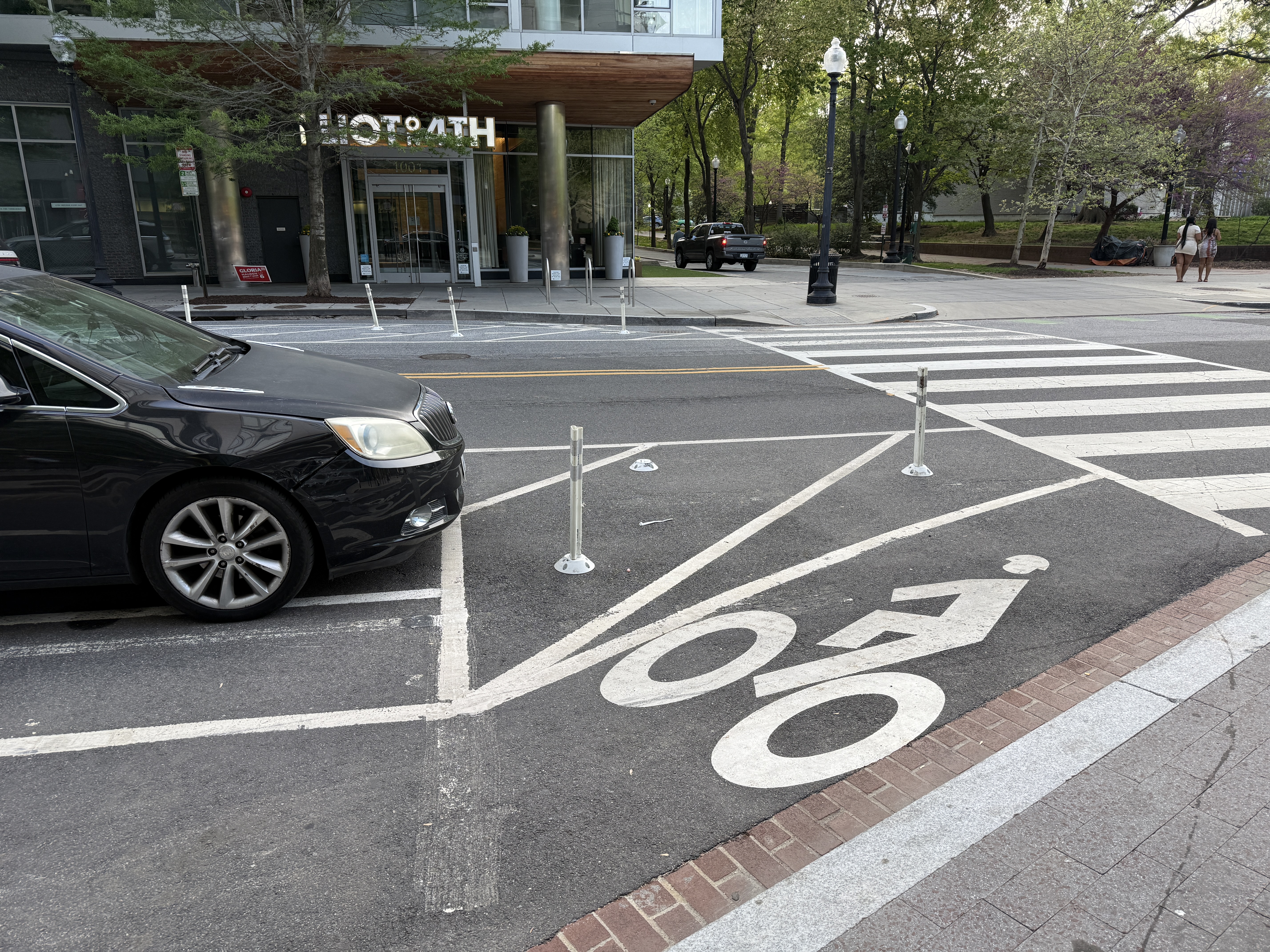 Two white sightline bollards installed at the 1001 4th St SW corner, but the parking-box striping ends flush with the bollards -- a parked vehicle sits right up to the daylighting buffer with no setback from the crosswalk.