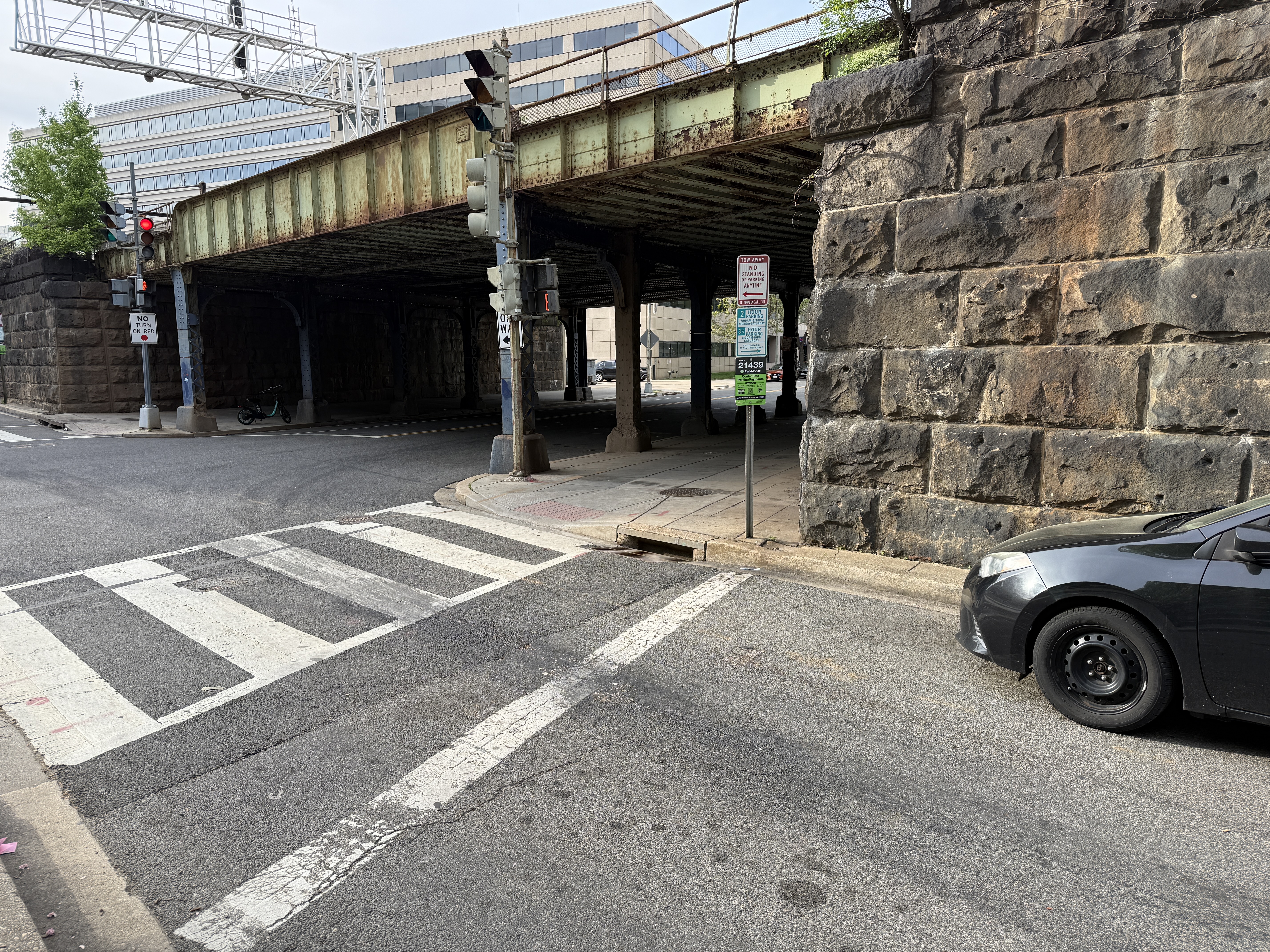 Dark sedan parked along the south curb of Virginia Avenue SW under the CSX rail bridge near 6th & Virginia. The vehicle's rear bumper is flush with the painted stop bar and well within the 40-foot intersection setback required by 18 DCMR 2405.2(c). The pole-mounted parking sign authorizes parking up to this location, so the violation is in DDOT's signage, not the driver's behavior.