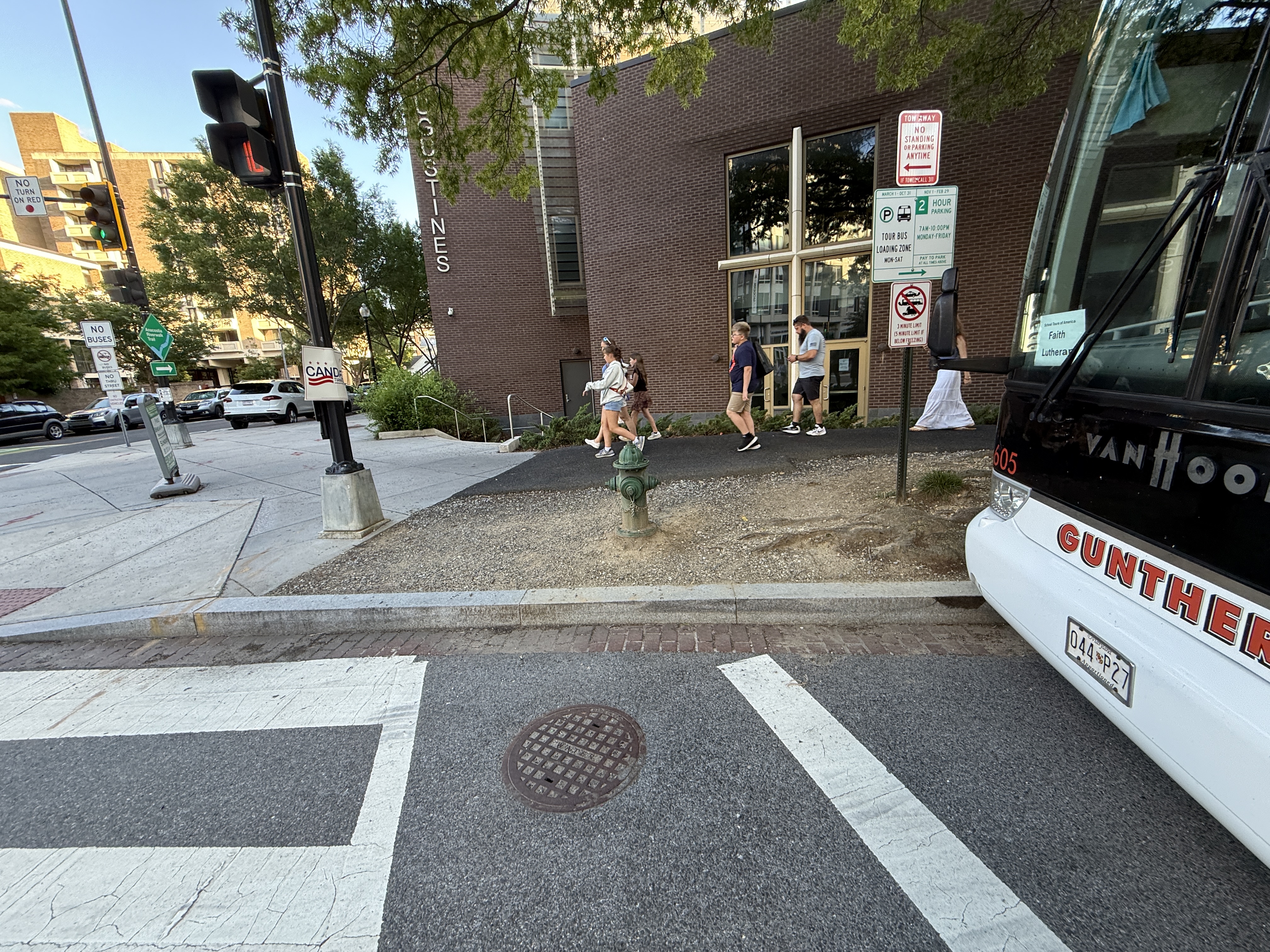 A 45-foot Van Hool coach bus parked in the Tour Bus Loading Zone at the NE corner of 6th St SW and M St SW, by St. Augustine's Episcopal Church. The loading-zone sign begins right at the pole next to the marked continental crosswalk, so the bus sits inside the daylighting setback and blocks pedestrian and driver sightlines. The violation is in DDOT's sign placement -- the bus is parked legally per the current sign.