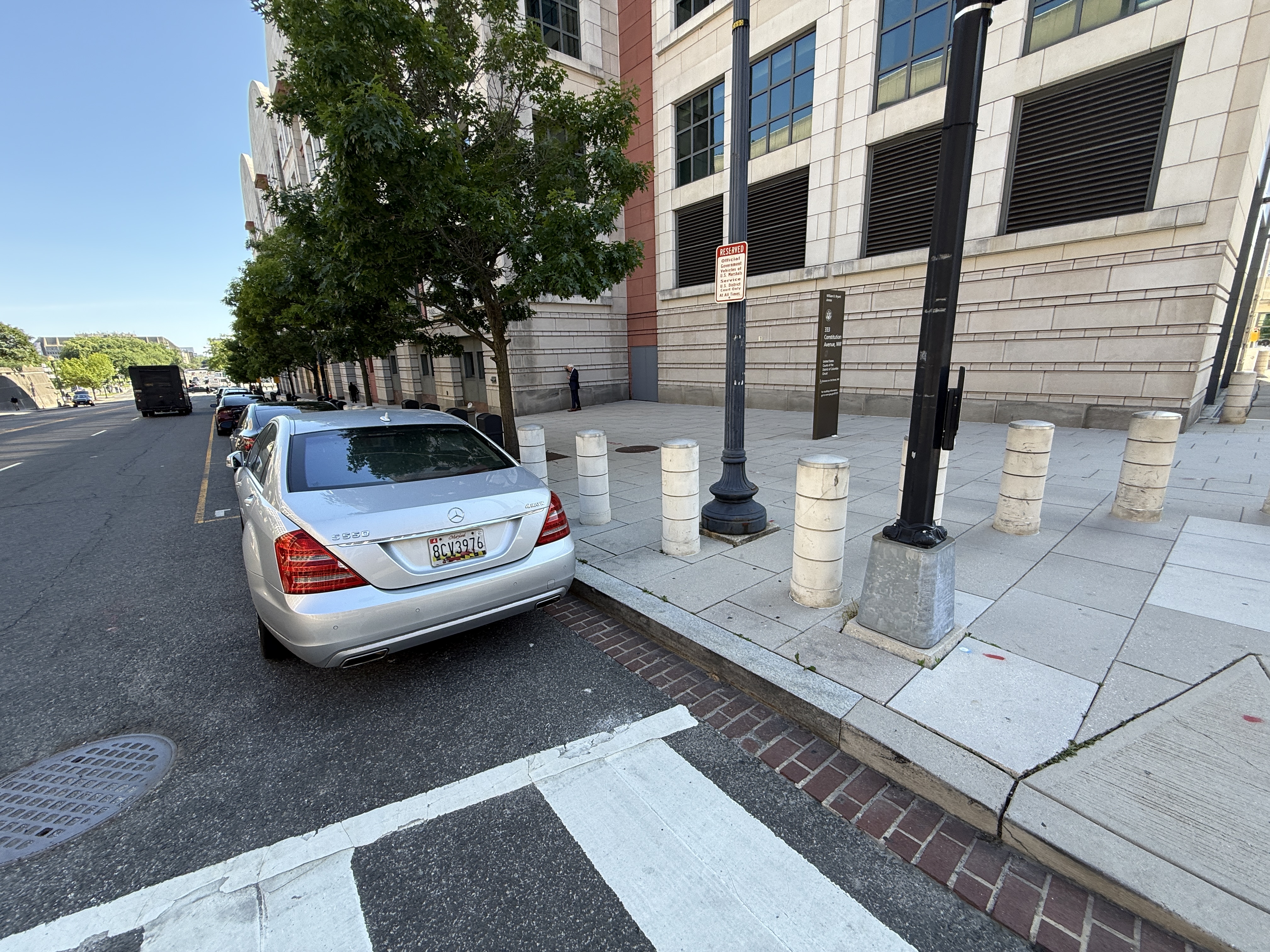 A private Maryland-tagged Mercedes S550 (MD plate 8CV3976) parked against the marked crosswalk at the corner of 333 Constitution Avenue NW and 3rd Street NW, beside a U.S. Marshals Reserved -- Official Government Vehicles At All Times sign that authorizes parking right up to the crosswalk at the federal courthouse.