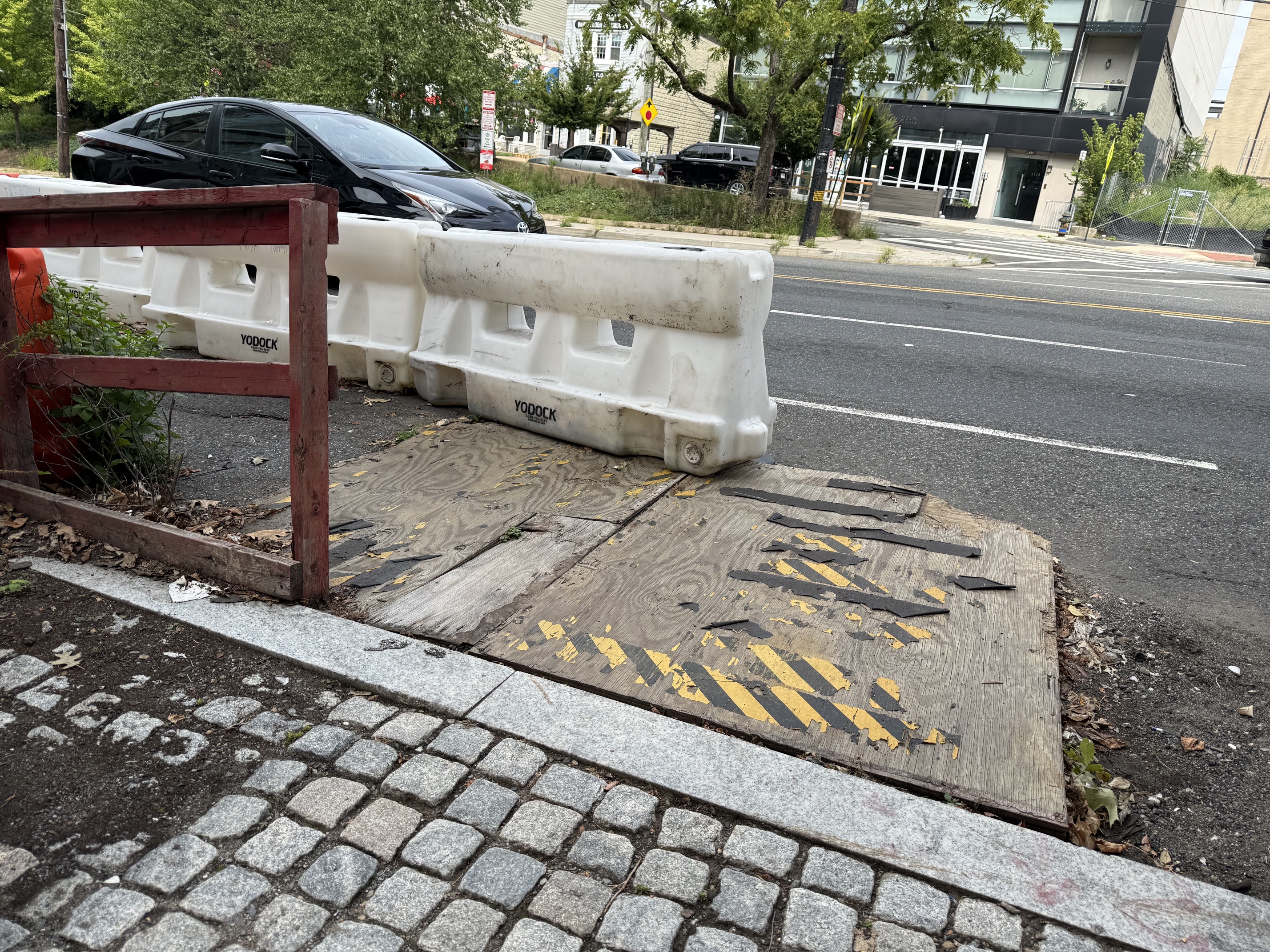 Close view of the plywood ramp surface: multiple warped sheets meeting at uneven joints, yellow and black anti-slip tape peeled off in strips, laid over the gutter against a line of white plastic barriers.