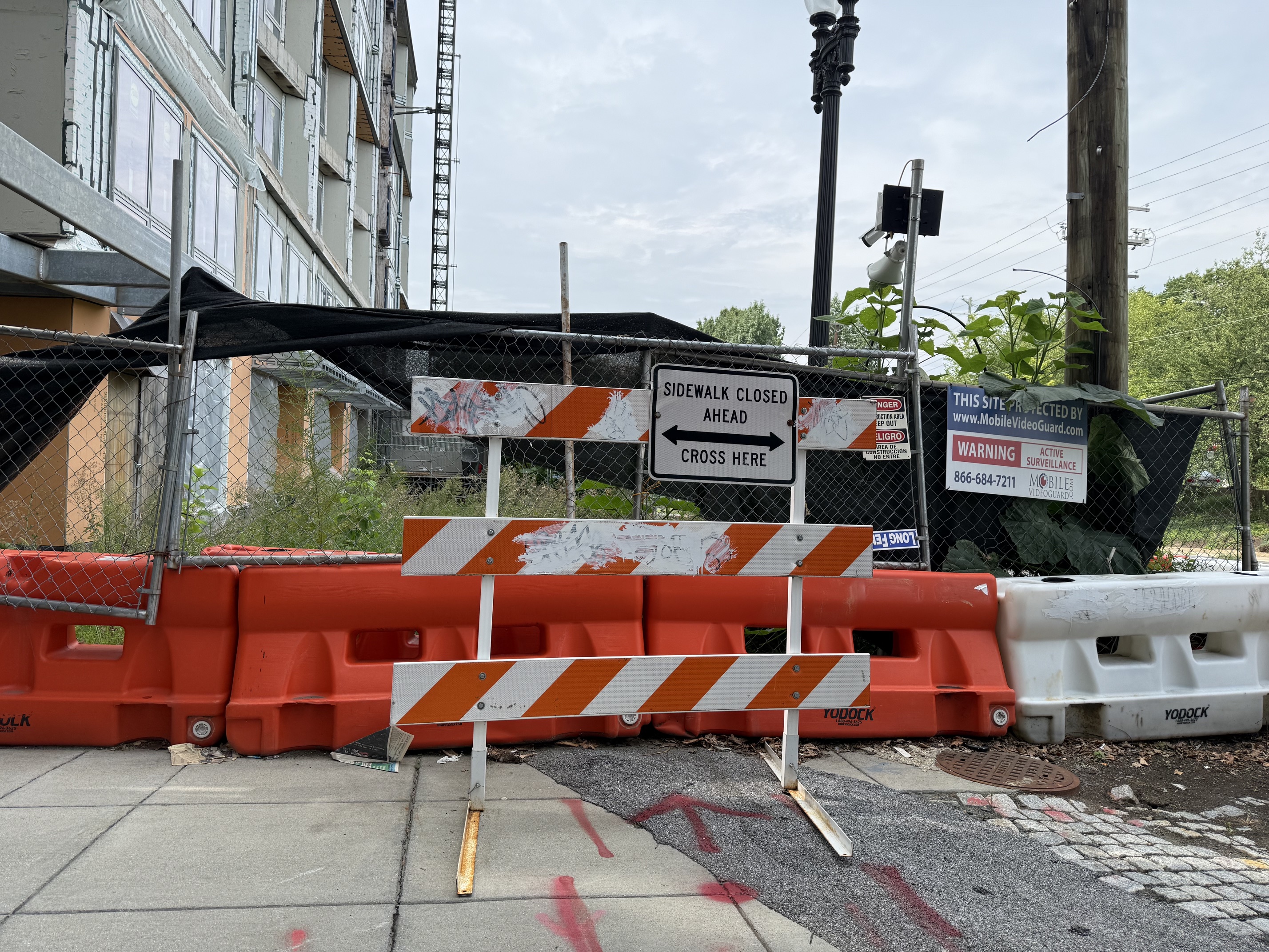 Weathered, graffitied orange-and-white barricades and a SIDEWALK CLOSED AHEAD, CROSS HERE sign in front of a chain-link fence draped in black mesh, with a stalled multi-storey building behind it.