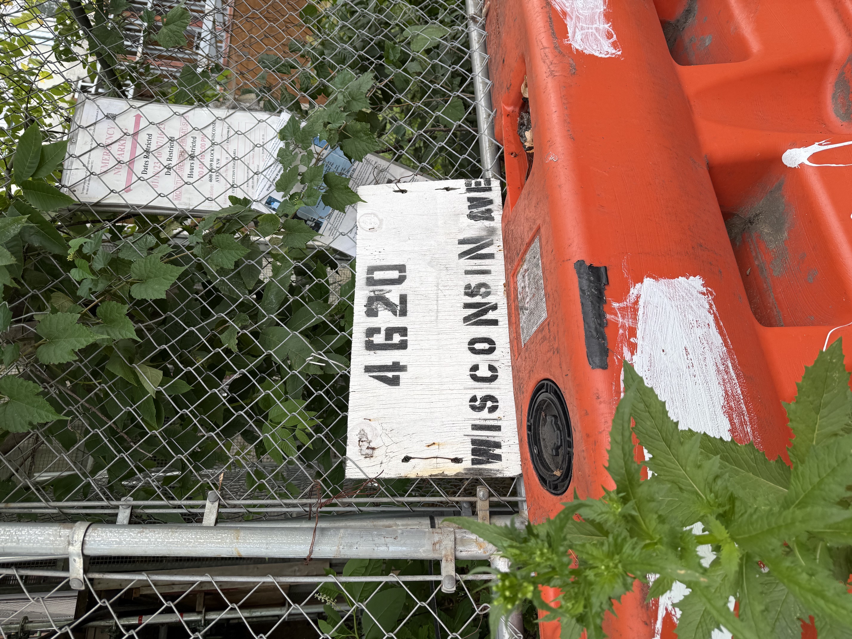 A hand-stenciled plywood placard reading 4620 WISCONSIN AVE, propped against an orange plastic barrier at the base of a chain-link fence, with the posted permit sign visible on the fence above it and weeds grown up around both.