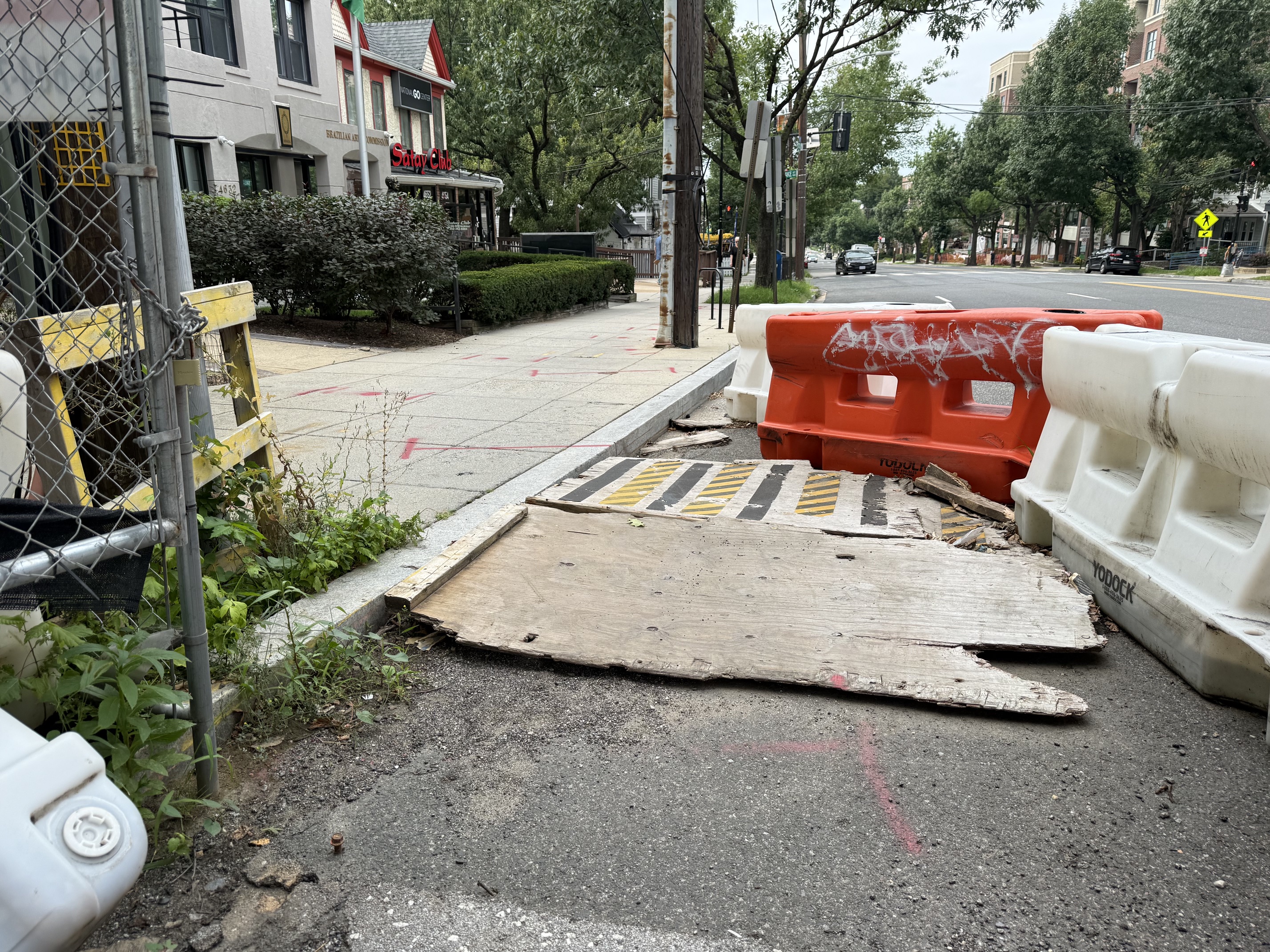 The temporary pedestrian ramp off the closed sidewalk at 4620 Wisconsin Avenue NW: a sheet of plywood, splintered and broken through at its edge, sagging off the curb into the roadway between orange and white Yodock barriers. The plywood is warped and uneven, the anti-slip tape has peeled away, and the broken edge leaves an unbridged drop -- there is no passable route here for a wheelchair, walker, or stroller. Behind it, SIDEWALK CLOSED AHEAD / CROSS HERE barricades close the block; the posted public-space permit on the fence expired July 16, 2023.