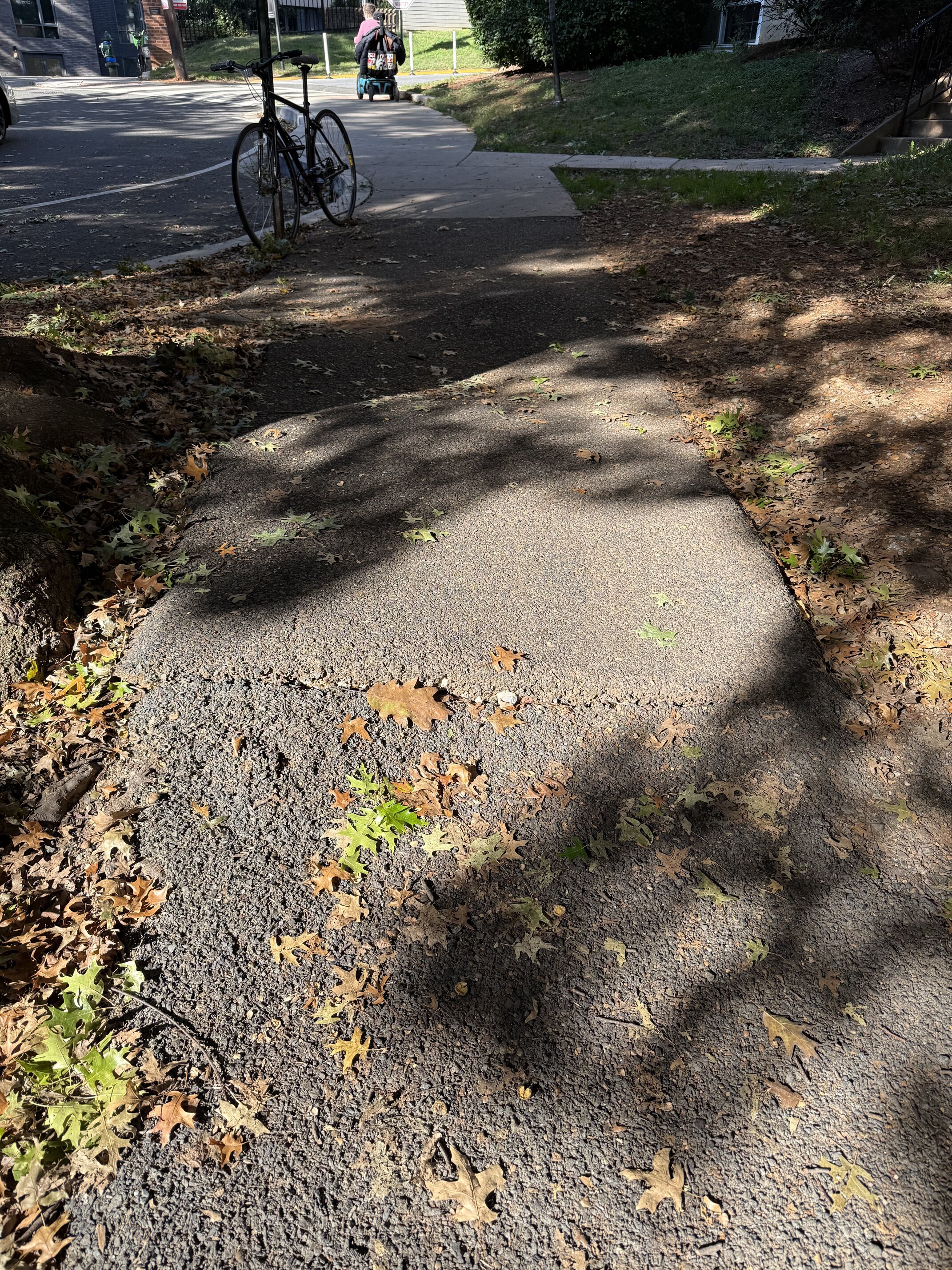 A stretch of sidewalk patched with asphalt, buckled into a raised hump with a cracked edge, covered in fallen leaves. Ahead, the sidewalk meets the street corner, where a person on a mobility scooter is moving along the sidewalk.