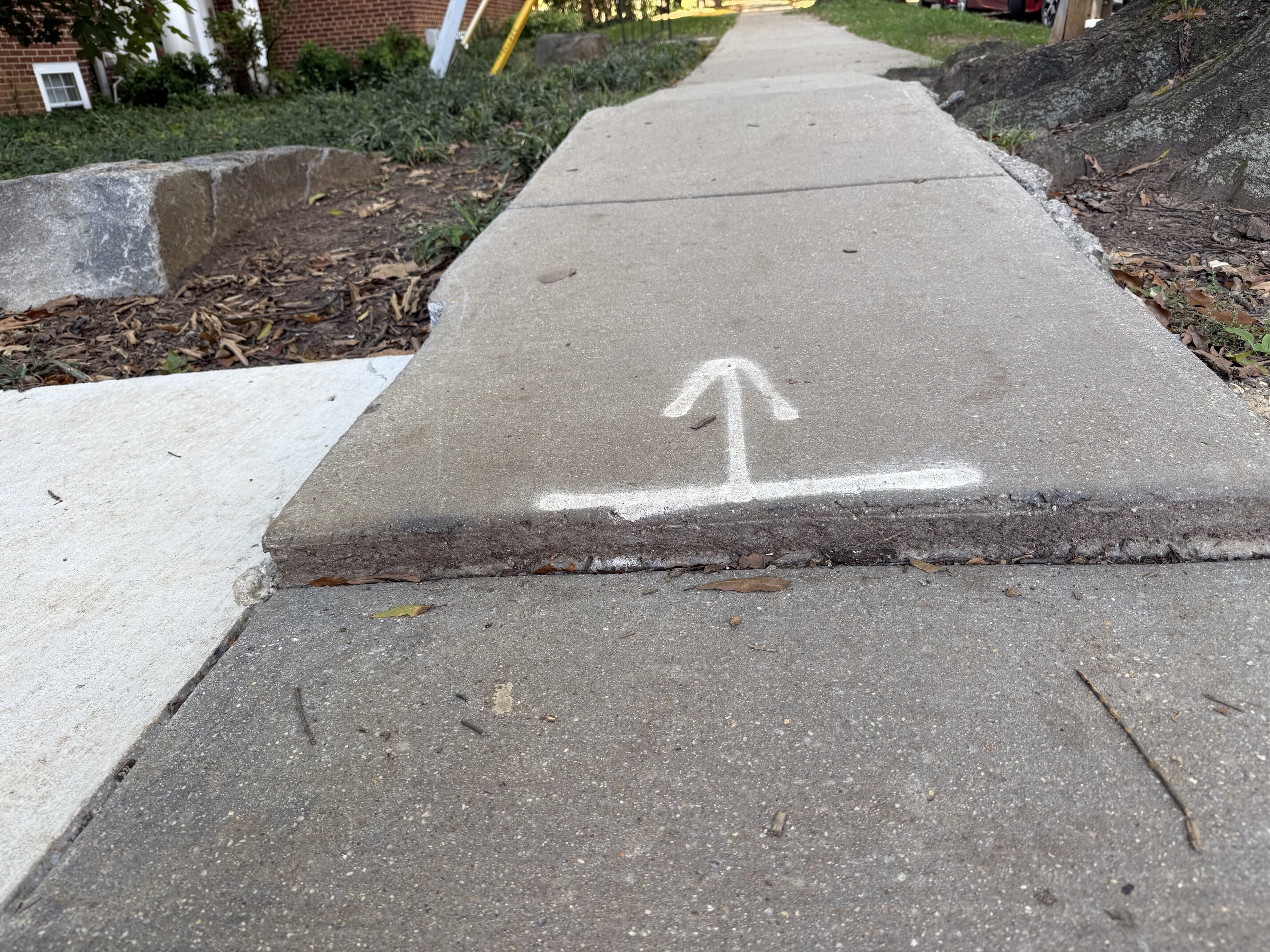 A concrete sidewalk slab on the 3700 block of 39th Street NW has heaved up above the slab next to it, leaving a raised, broken-edged lip across the full width of the walk. Someone has spray-painted a white arrow and line on the raised slab to mark the trip edge. A second frame on the same block shows an asphalt-patched stretch of sidewalk buckled into a raised hump with a cracked edge, with a person on a mobility scooter on the sidewalk ahead.